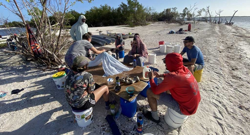 People gather around a makeshift table that is set up on buckets. They are on a sandy beach. 